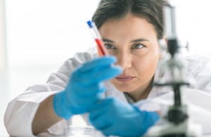 <p>A female laboratory technician examines blood vial sample. Medical gloves and a lab coat are worn for protection. Focus is on the laboratory technician.</p>